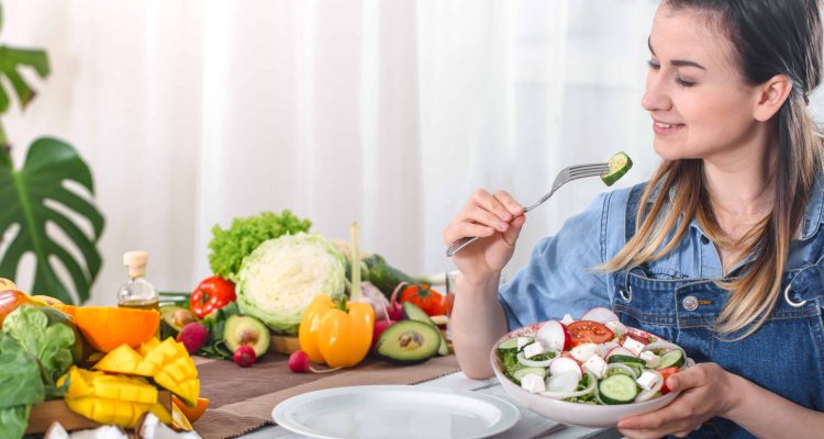 young-happy-woman-eating-salad-with-organic-vegetables-table-light-background-denim-clothes-concept-healthy-home-made-food-scaled